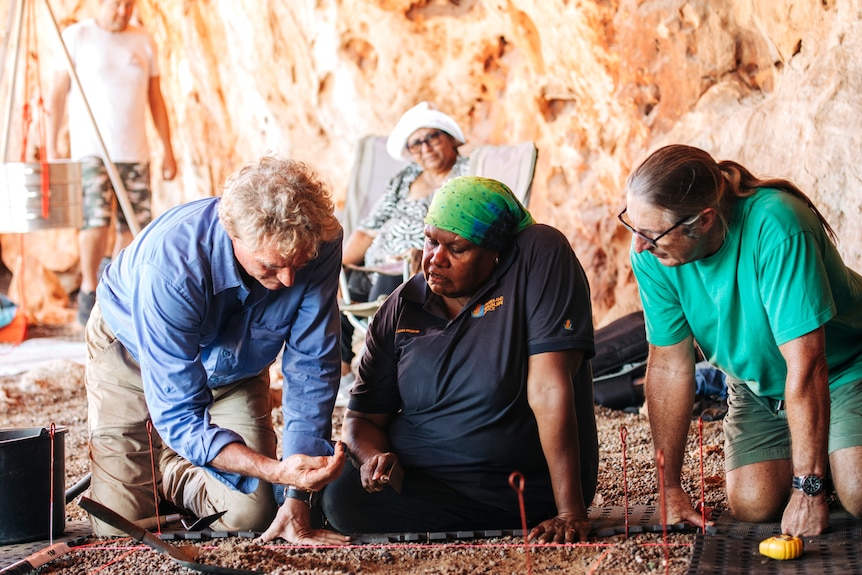 In a brightly lit rock shelter, a woman and two men sit and kneel over looking at findings from an archaelogical dig.