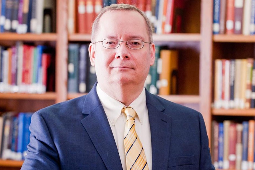 A man in a suit with glasses sits in front of a bookshelf.