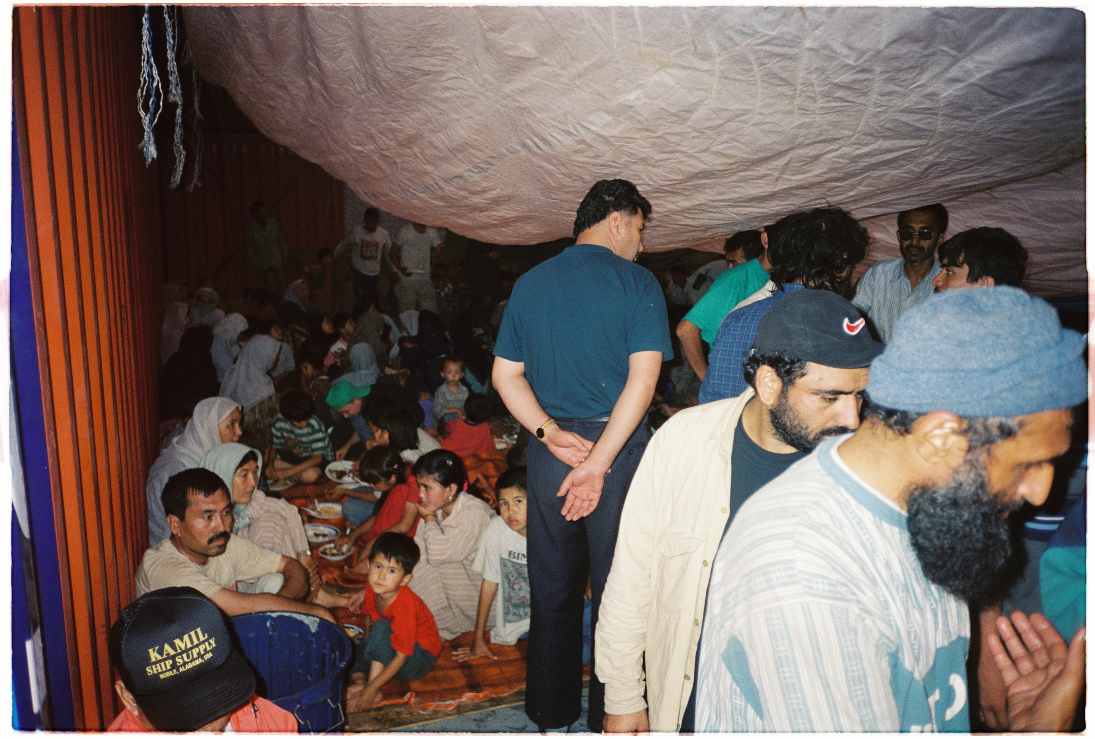 A crowd of people, including young children, stand and sit on the floor beneath a tarp.