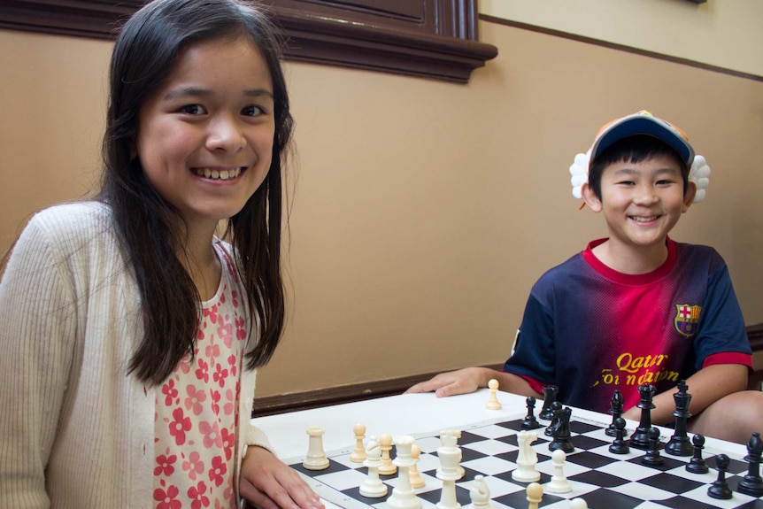 A young girl and boy sit on either side of a chess board.