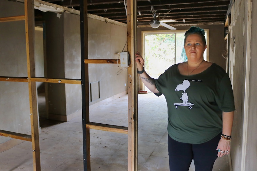 An image of Terri Thompson standing in her home with wooden frame of wall visible, flood covered in mud and damaged walls