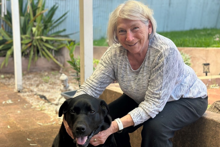 A woman with short grey hair sitting down and smiling next to her black dog. 