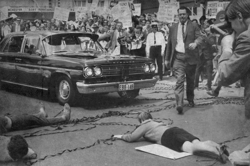 Protesters lie on the road in front of a limousine, in front of a large crowd waving signs.