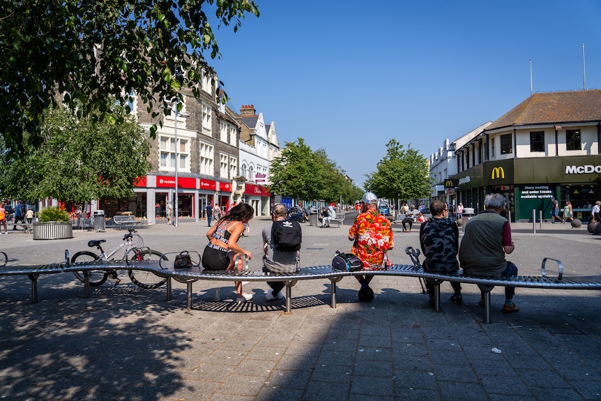 Five people sit on a steel public bench in the sun in a street with trees providing some shade on a sunny day