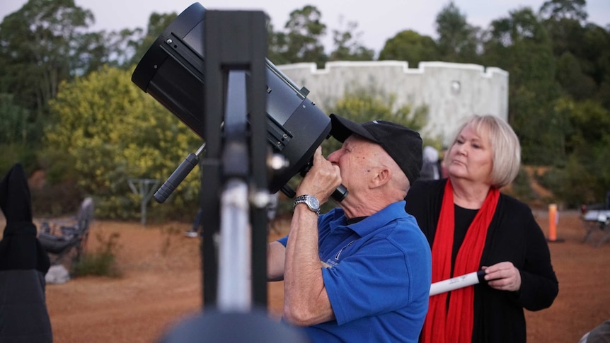 And older man looks at the sky through a large telescope next to a woman, while standing outside on red earth and scrub.