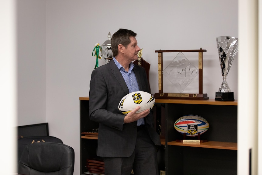 Man in suit stands with a rugby league football in his hands while looking at trophies.