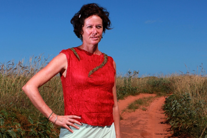 A woman with short brown hair wears a red shirt and stands in the red dirt with one hand on her hip