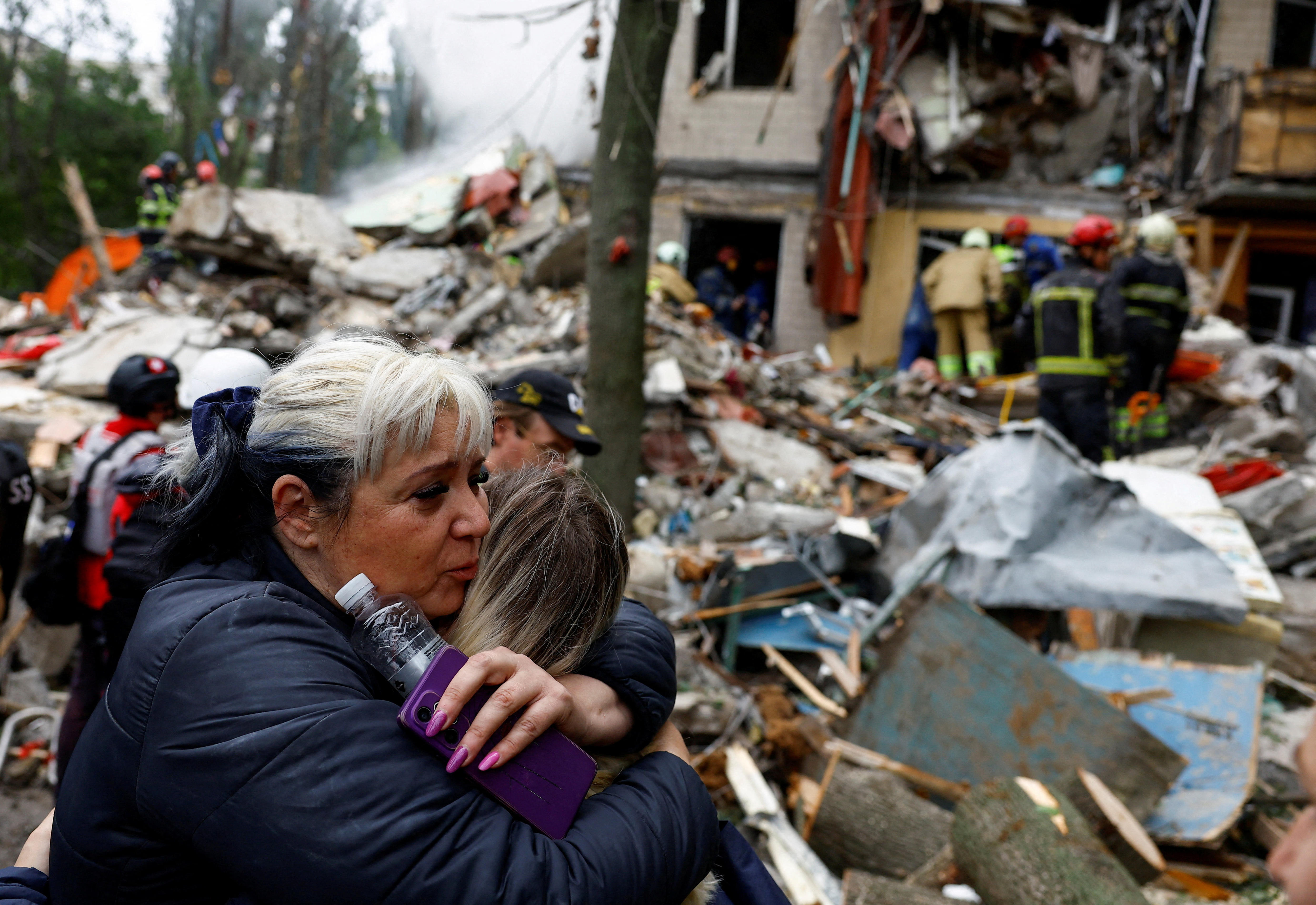 A woman with black and white hair hugging another female, while holding a water bottle and mobile phone, in front of debris
