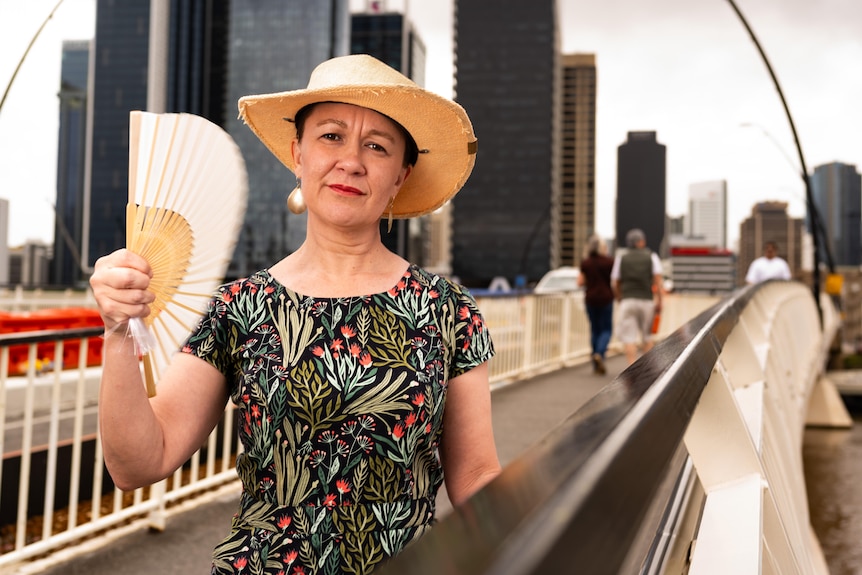 A woman in a hat cools herself with a fan while standing on a bridge in a CBD.