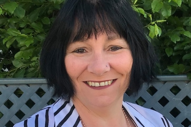 A middle-aged woman with black hair smiles at the camera. Her shirt is purple with black stripes.