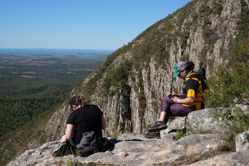 Two women sit on some rocks, sheer cliff face with shrubs, land and ocean in background.