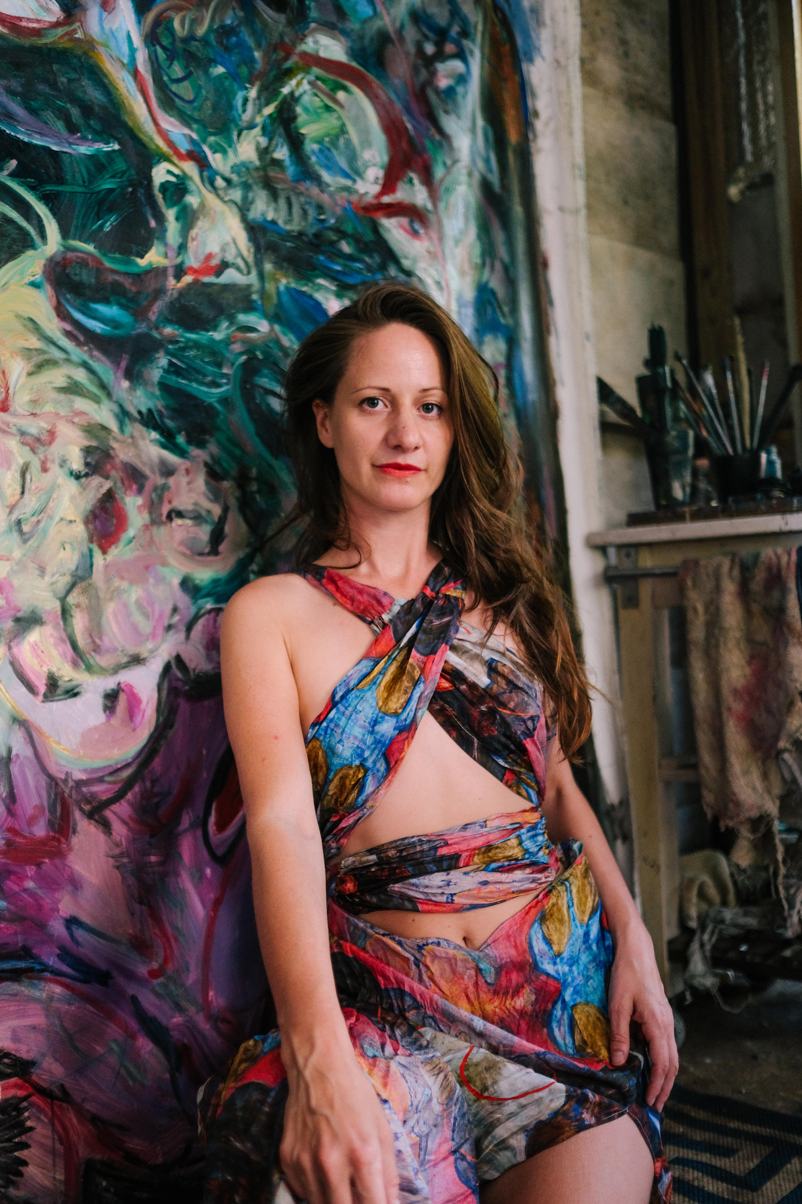 Walker in her studio, looking strong at the camera. She kneels in front of a painting, wearing a colourful sarong dress.