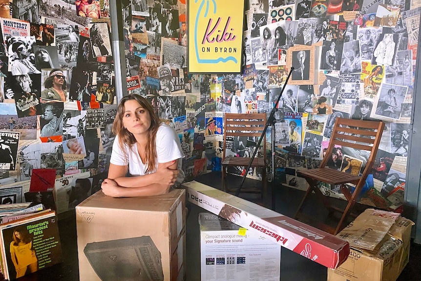 A young woman leans on moving boxes inside a venue wallpapered with band posters.
