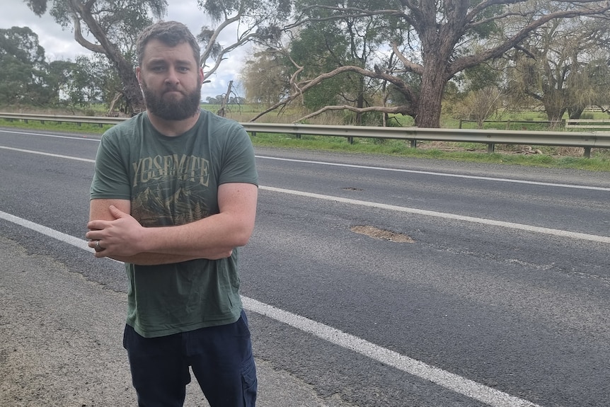 A man stands with his arms folded alongside a road with a pothole on it.