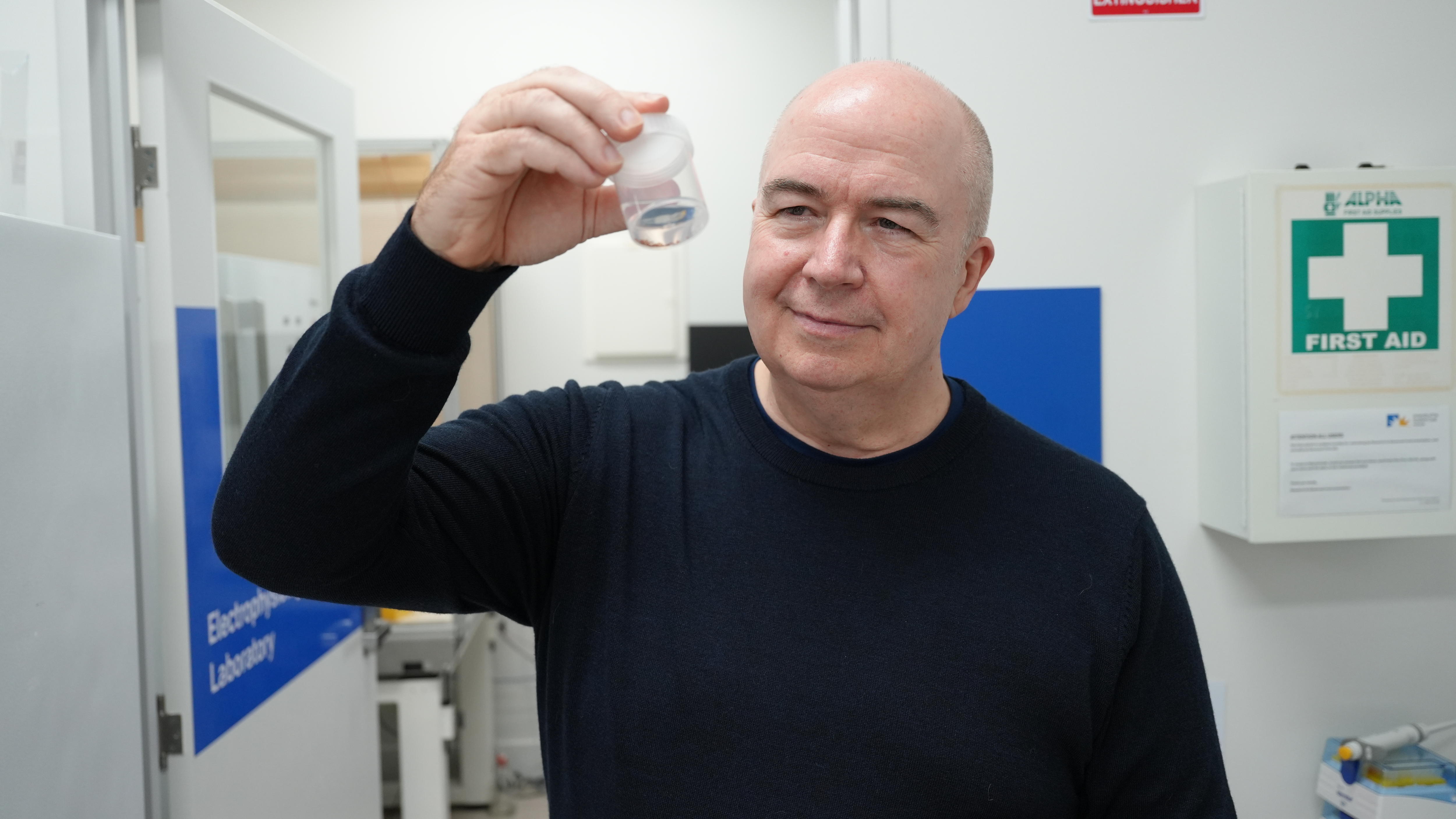 A midddle-aged man in a dark jumper holds up a sample container while standing in a laboratory.