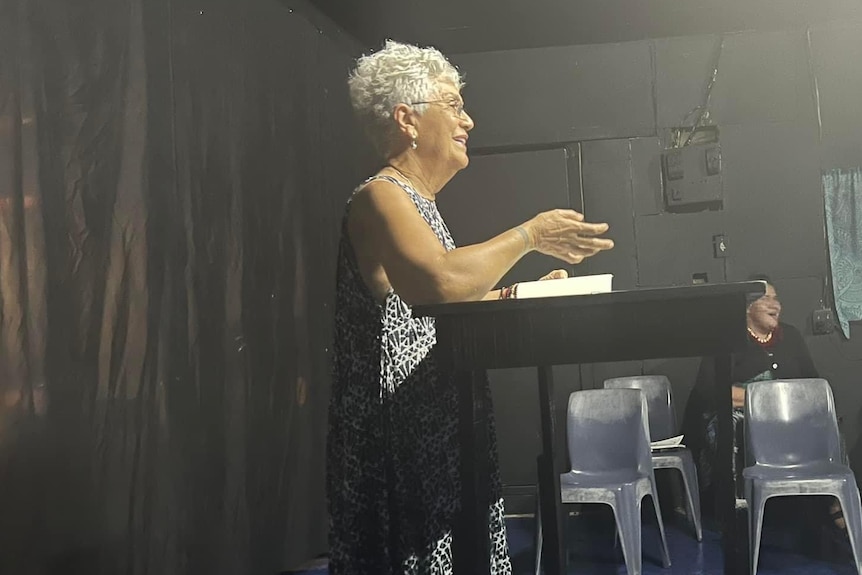 An older Samoan woman, standing at a table and speaking.