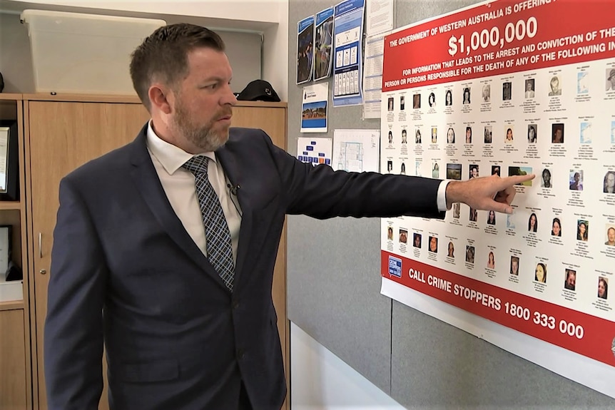 A man in a suit stands in an office pointing at a board advertising rewards for information about crimes