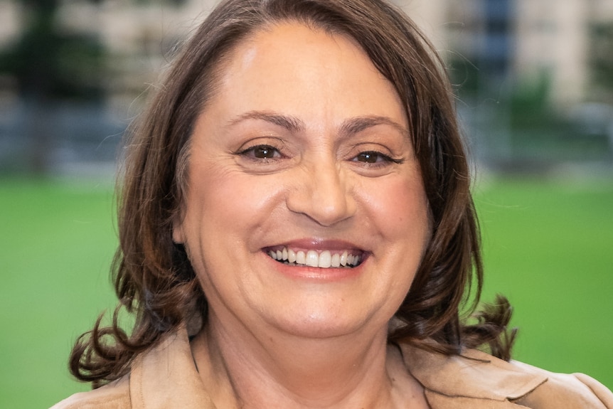 A woman with brown hair smiles wearing a light brown coat