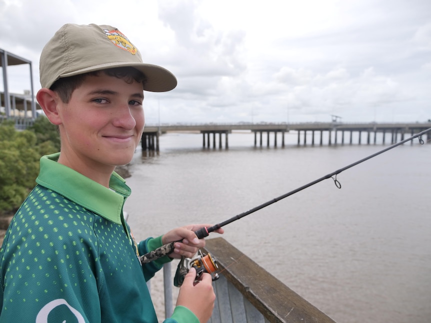 A teenager in a cap stands with a fishing line in his hand, a bridge is visible in the background.
