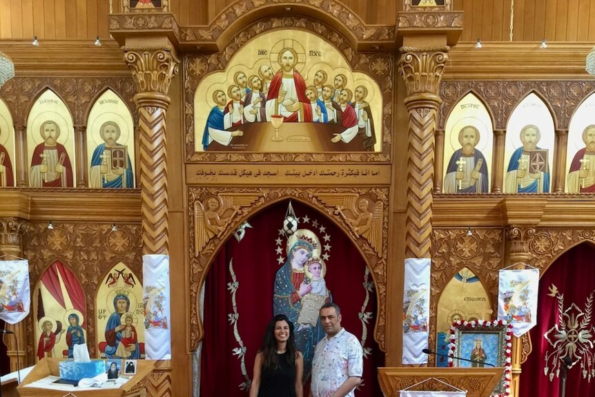 A man and woman stand in front of an ornate wall filled with religious art.