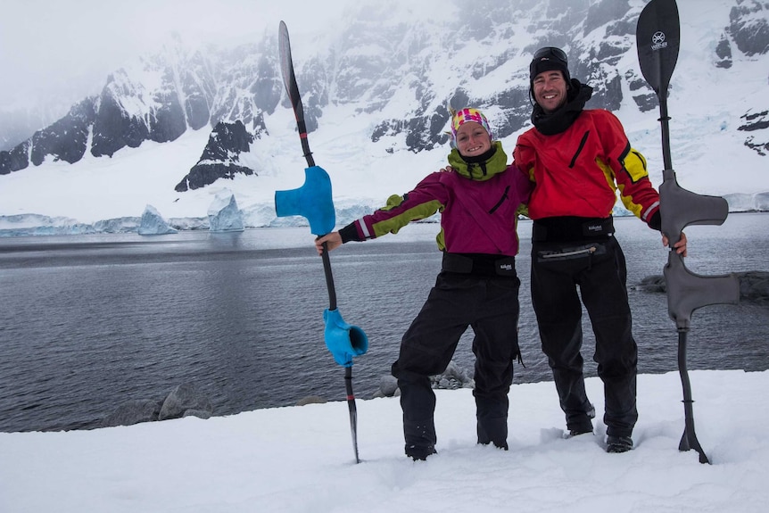 Sophie Ballagh and Ewan Blyth smile and hold their kayak paddles as they stand on ice by Antarctic waters.