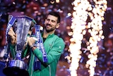 Novak Djokovic holds a trophy aloft after winning a tennis tournament, as fireworks go off in the background.
