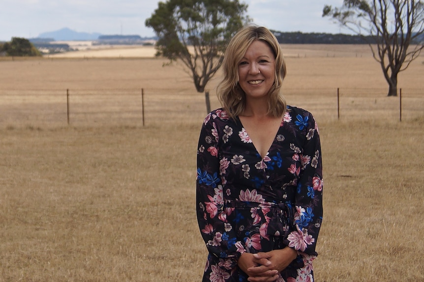 Woman with long blonde hair, wearing long sleeved floral shirt, smiling, standing in barron farmland