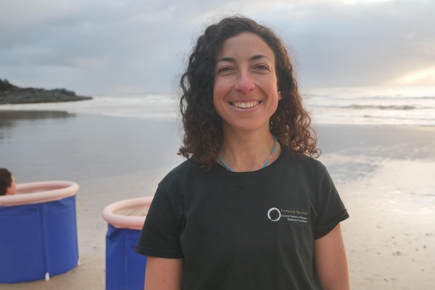 A woman in a black t-shirt smiles happily on a beach in front of some portable baths