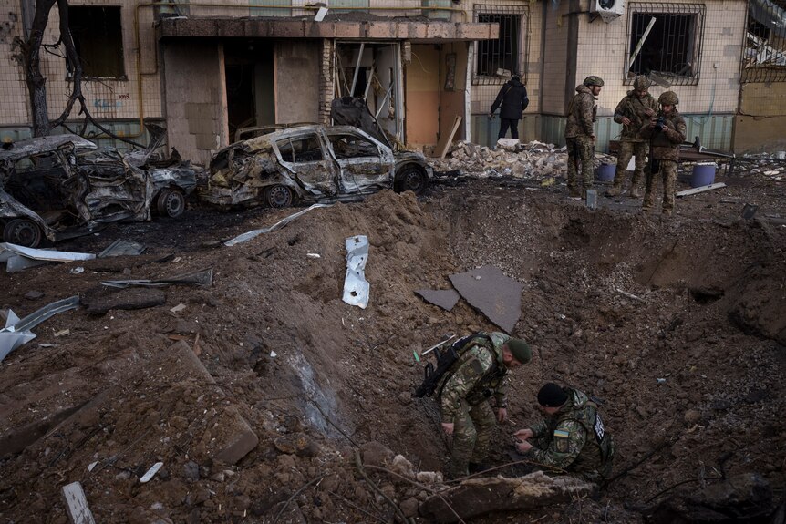 Two soldiers inspect a crater at the site of an explosion after bombing while three others look on.