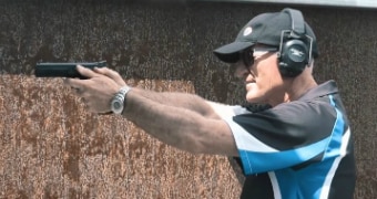 Steve Dickson, wearing a hat, sunglasses and ear protectors, poses with a gun in a firing range.