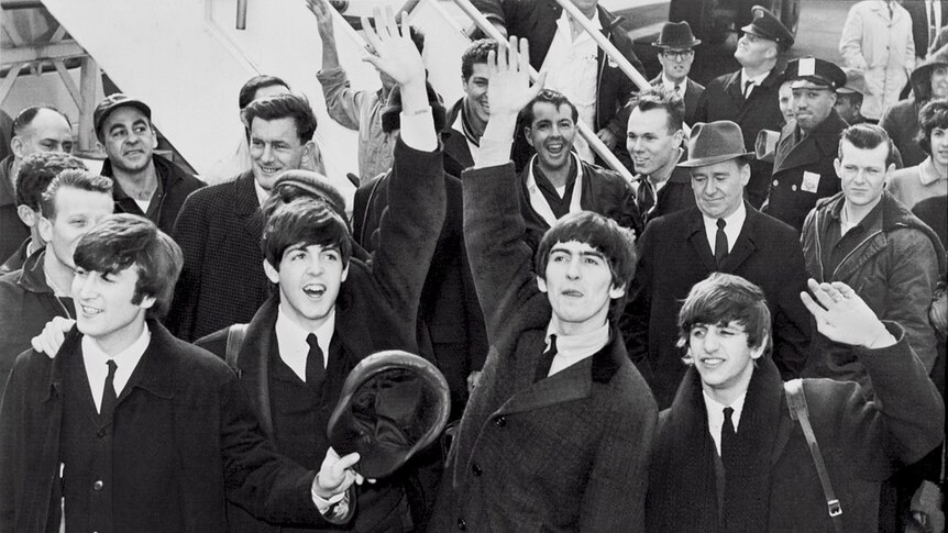 Black and white picture of The Beatles getting off a plane, surrounded by people.