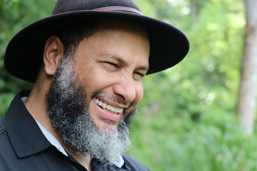 A bearded man wearing a black wide-brimmed hat smiles a big smile.