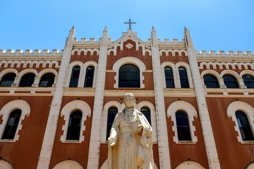 Statue outside a large monastery white and dusty orange building with cross on top of building.