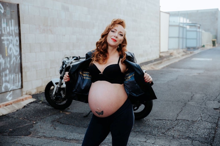 A heavily pregnant woman with red hair, wearing motorcycle leathers.