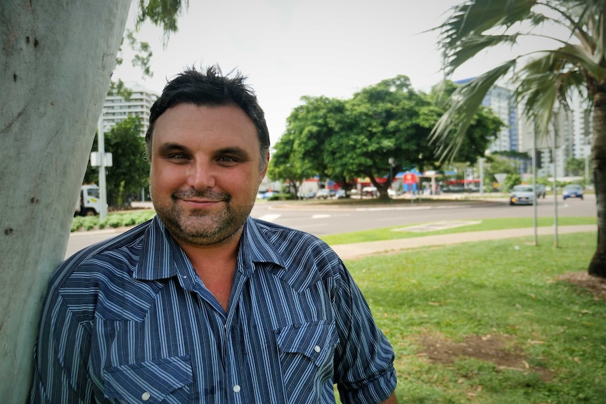 Man leaning against a ghost gum with a flourishing tree on a roundabout behind.