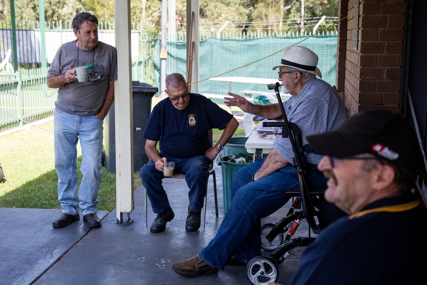 a man standing up leaning against a pole and holding a mug with three men sitting on chairs under a pergola