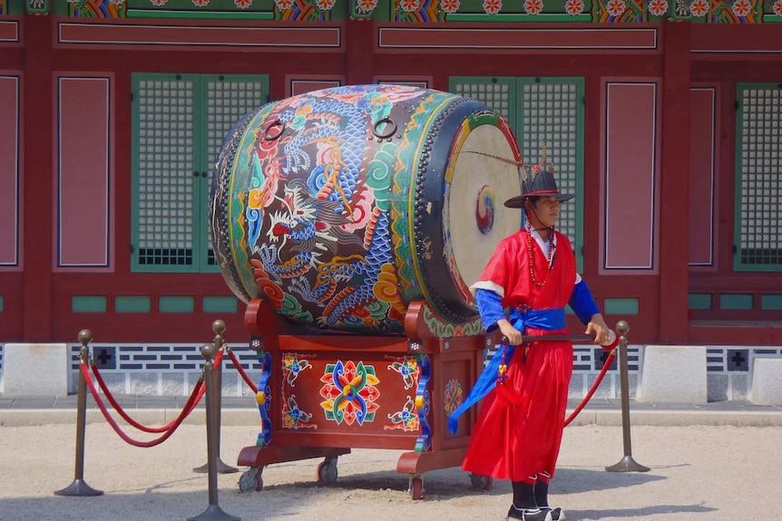 Man in Korean ceremonial robes of red and blue standing in front of large, ornately decorated drum.