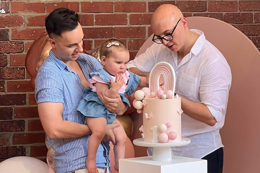 Two dads celebrate their daughter's first birthday party; one holds the baby while the other cuts a pale pink cake.
