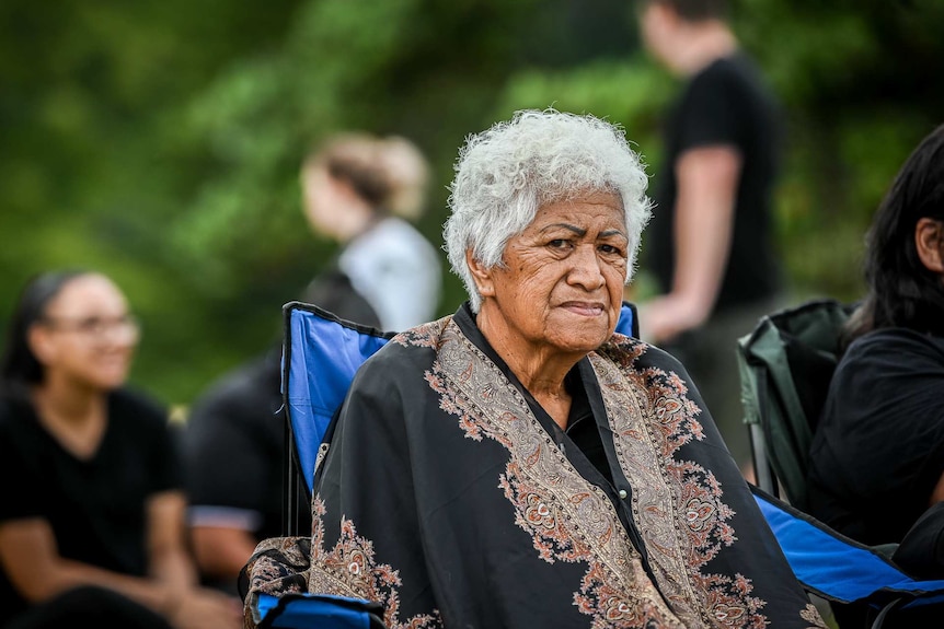 An older woman wearing a black top sits in a chair and looks pensive