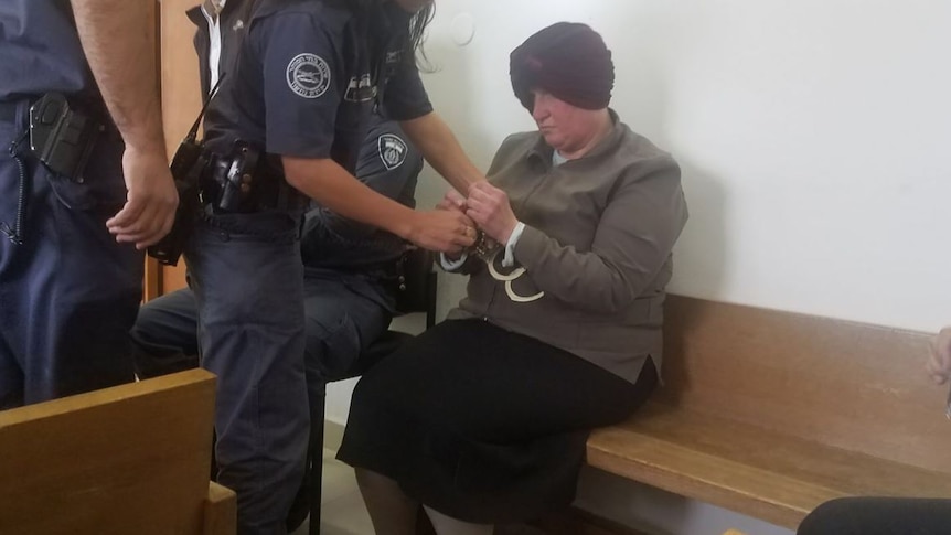Malka Leifer sits on a wooden seat as a police officer handcuffs her.