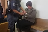 Malka Leifer sits on a wooden seat as a police officer handcuffs her.