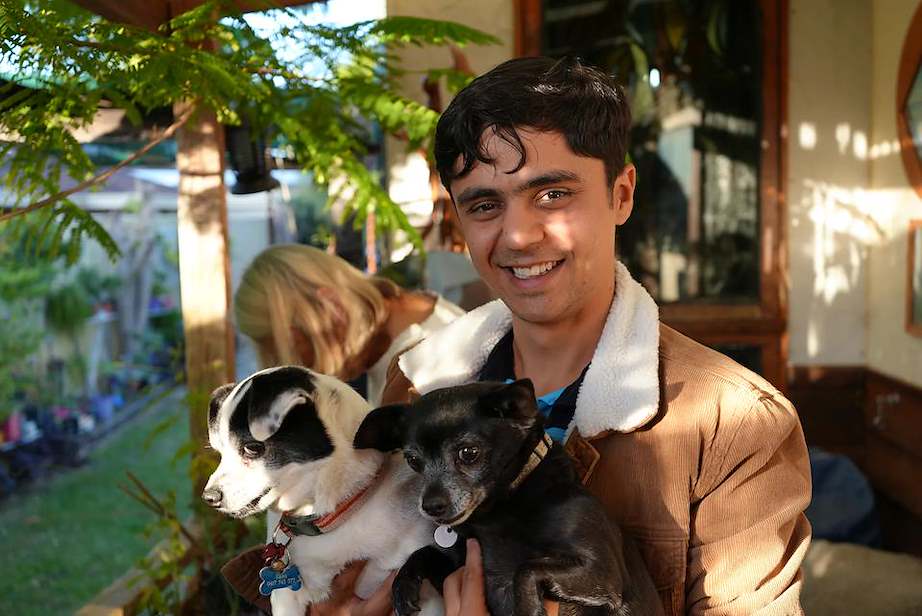 A man with dark hair wearing a camel jacket holding two small dogs, smiling at the camera, stood outside.