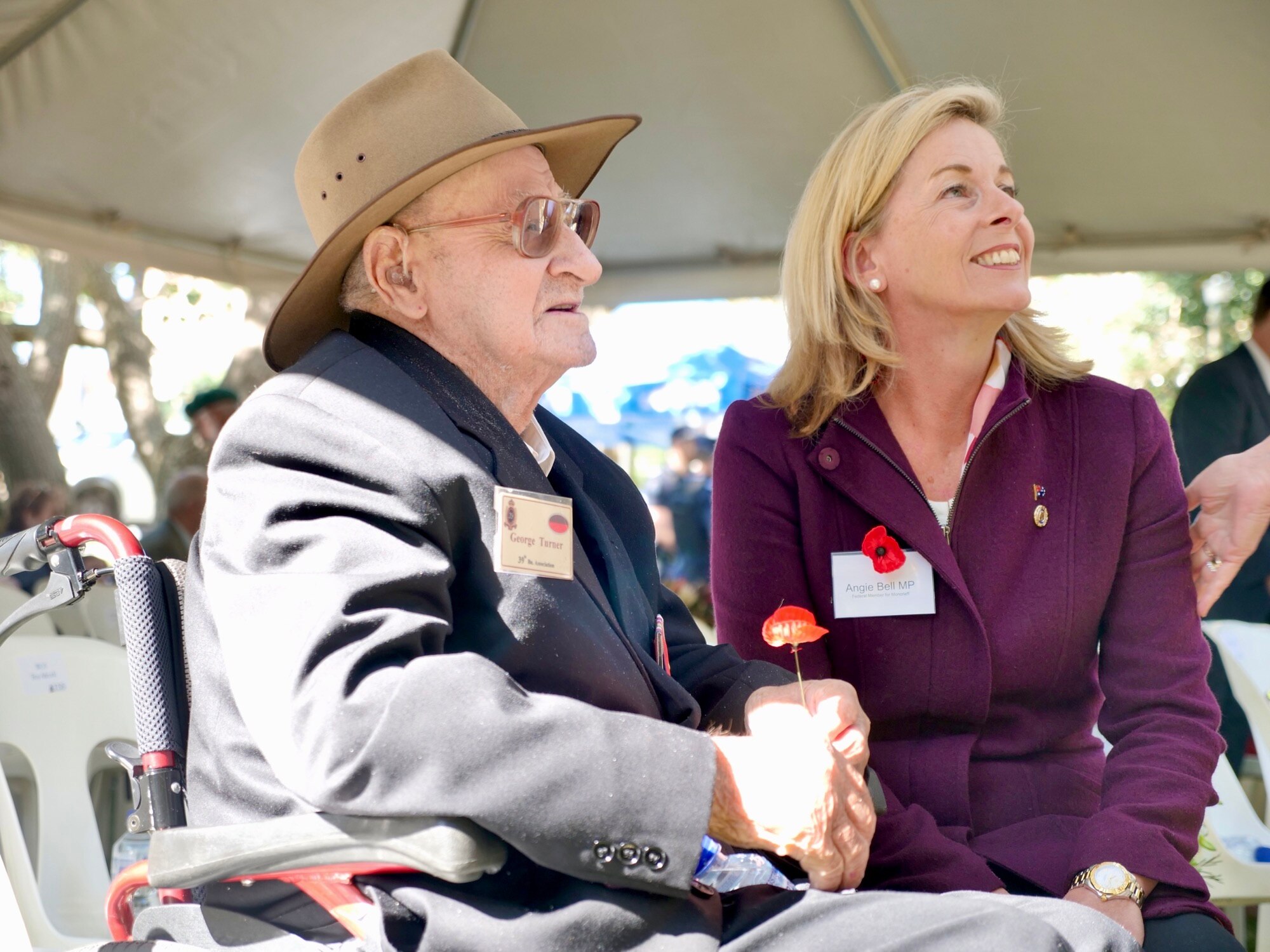 an elderly man in a wheelchair sits beside a smiling younger woman in a marquee.