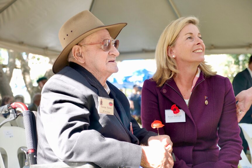 an elderly man in a wheelchair sits beside a smiling younger woman in a marquee.