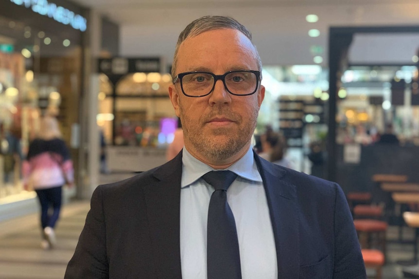 A middle aged man in a suit and tie stands posing for a photo in a shopping centre.