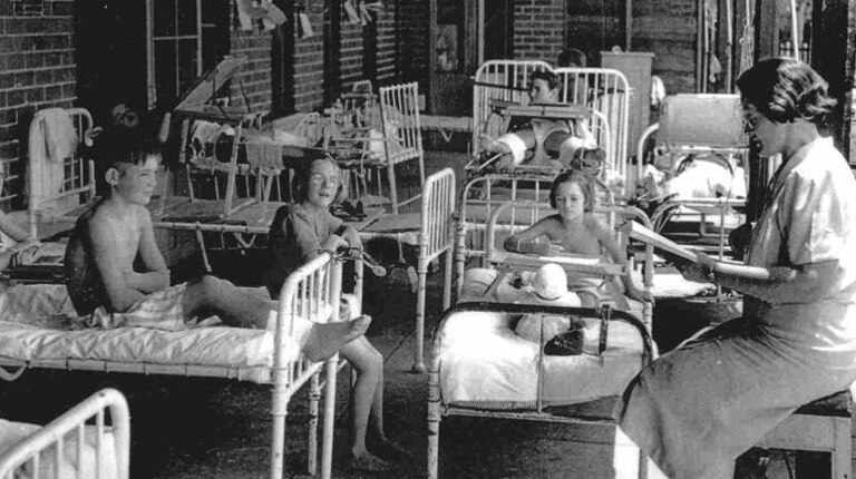 Black and white photo of children sitting on hospital beds with teacher reading from book