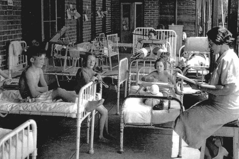 Black and white photo of children sitting on hospital beds with teacher reading from book