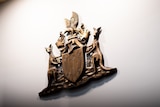A bronze Commonwealth Coat of Arms hangs on a white wall inside a court house. I