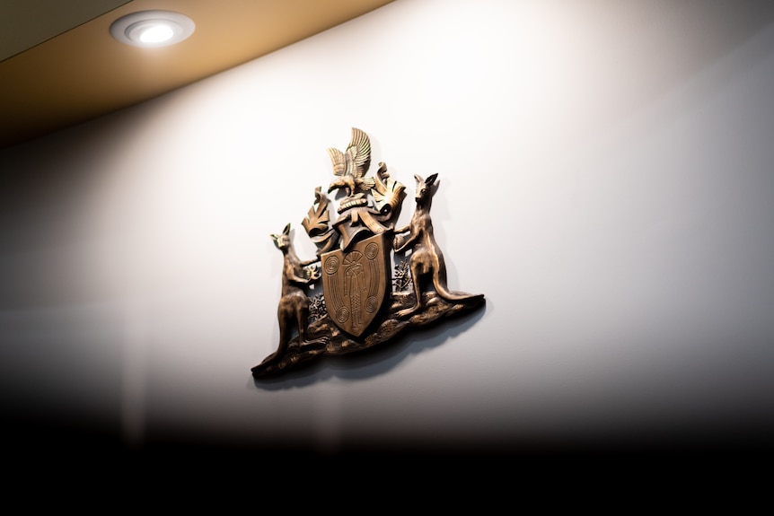 A bronze Commonwealth coat of arms hangs on a white wall inside a courthouse.  I