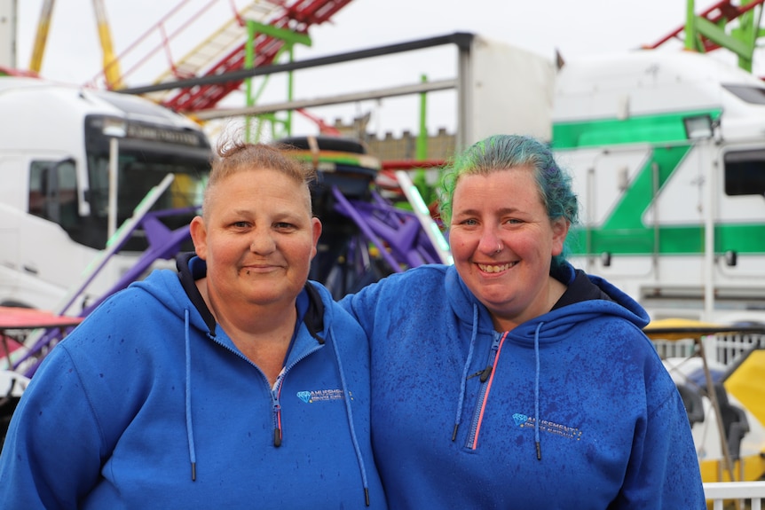Two woman in blue jumpers stand in front of show rides smiling. 